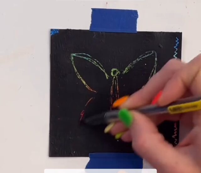 Cartes à gratter, troisième étape : creuser un papillon dans la couche noire avec un bâtonnet en bois, les couleurs réapparaissent sous le trait